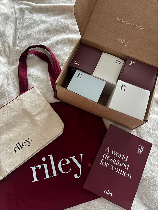 Riley brand gift set arranged on a white fabric surface, including an open brown box with multiple small branded boxes, a cream tote bag, a deep burgundy pouch, and a matching printed card reading 'A world designed for women'.