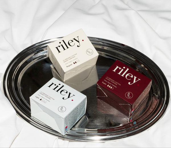 Three small Riley product boxes in cream, white, and deep red are arranged on a round silver tray over a white fabric background.