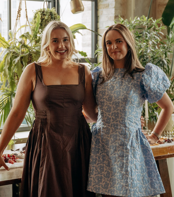 The two co-founders of Riley standing indoors in a bright, plant-filled space. The woman on the left wears a dark brown sleeveless dress, and the woman on the right wears a light blue puff-sleeve dress with a beige abstract pattern.