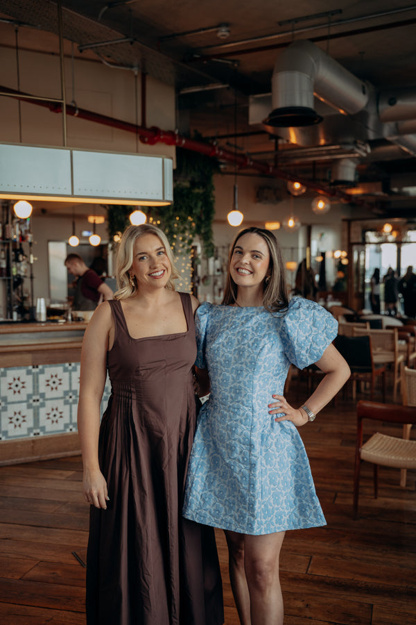 The two female co-founders of Riley, Fiona Parfrey and Aine Kilkenny posing together in a warm, industrial-style cafe interior. The woman on the left wears a sleeveless brown dress, and the woman on the right wears a light blue and white patterned puff-sleeve mini dress.