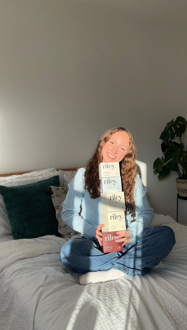 A woman sitting cross-legged on a bed holding a stack of four boxes of Riley period care in a softly lit bedroom with pillows and a plant in the background.