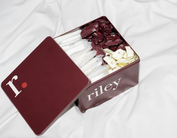 An open dark red metal box of Riley tin, showing tampons and pads inside, set on a white fabric background.
