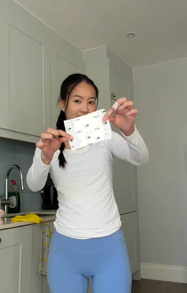 A woman in a white long-sleeve top and blue shorts stands in a kitchen holding up a riley pad.