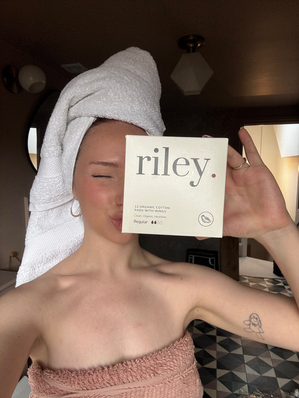 A woman wrapped in a white towel on her head and a dusty pink towel on her torso holds up a cream-colored Riley period care box in front of her face in a bathroom setting.