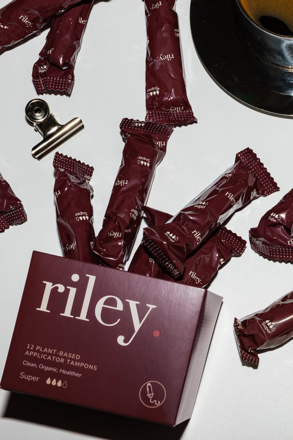 Riley super tampons shown with their branded box on a white desk surface, surrounded by coffee cups, eyeglasses, binder clips, and a pen.