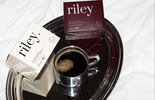 Two boxes of Riley tampons, one white and one dark burgundy, arranged on a oval silver tray next to a small cup of black coffee on a white fabric background.