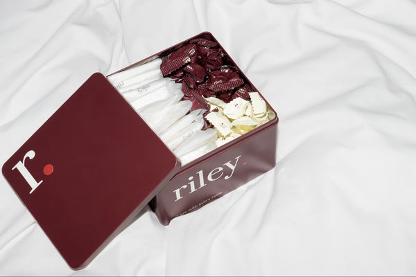 An open dark red metal box of Riley tin, showing tampons and pads inside, set on a white fabric background.